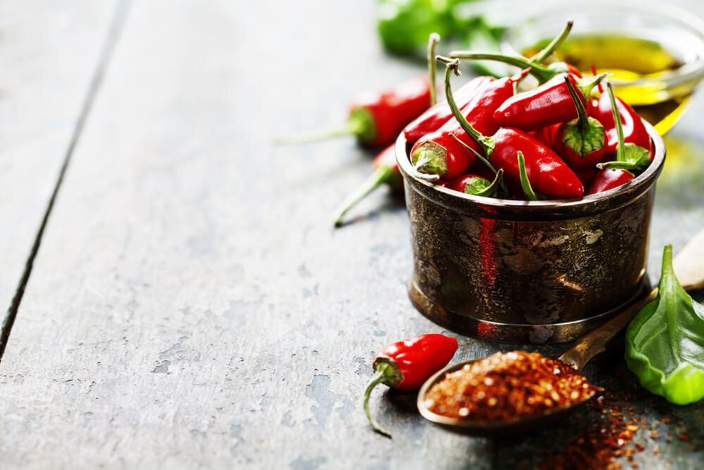 Freshly picked red chili peppers in a metallic bowl, and next to it are herbs and spices on a spoon.