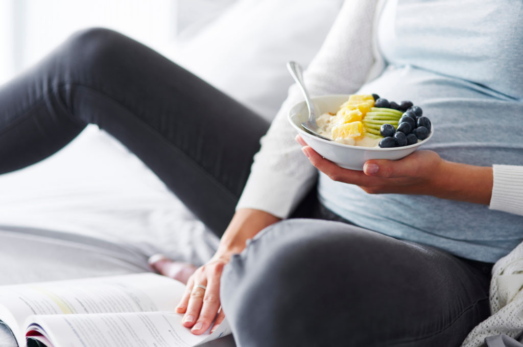 A pregnant woman is in bed. She’s having a healthy breakfast and reading a book.