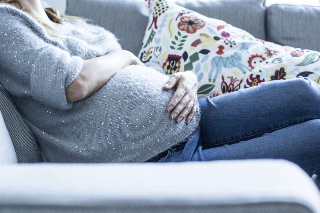 Eine junge schwangere Frau sitzt auf der Couch und streichelt ihren Bauch.