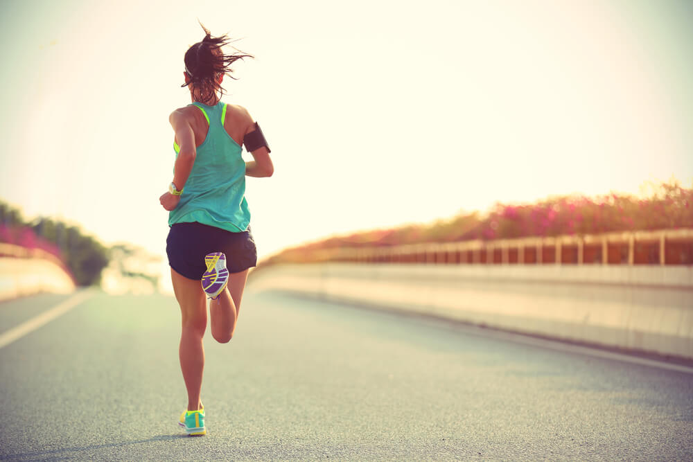 A female runner wearing sports clothes is running on an asphalt road towards the sunset.
