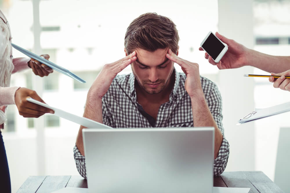 Мужчина на работе сидит перед компьютером, обхватив голову руками, потому что у него сильный стресс, так как на него давят со всех сторон. 