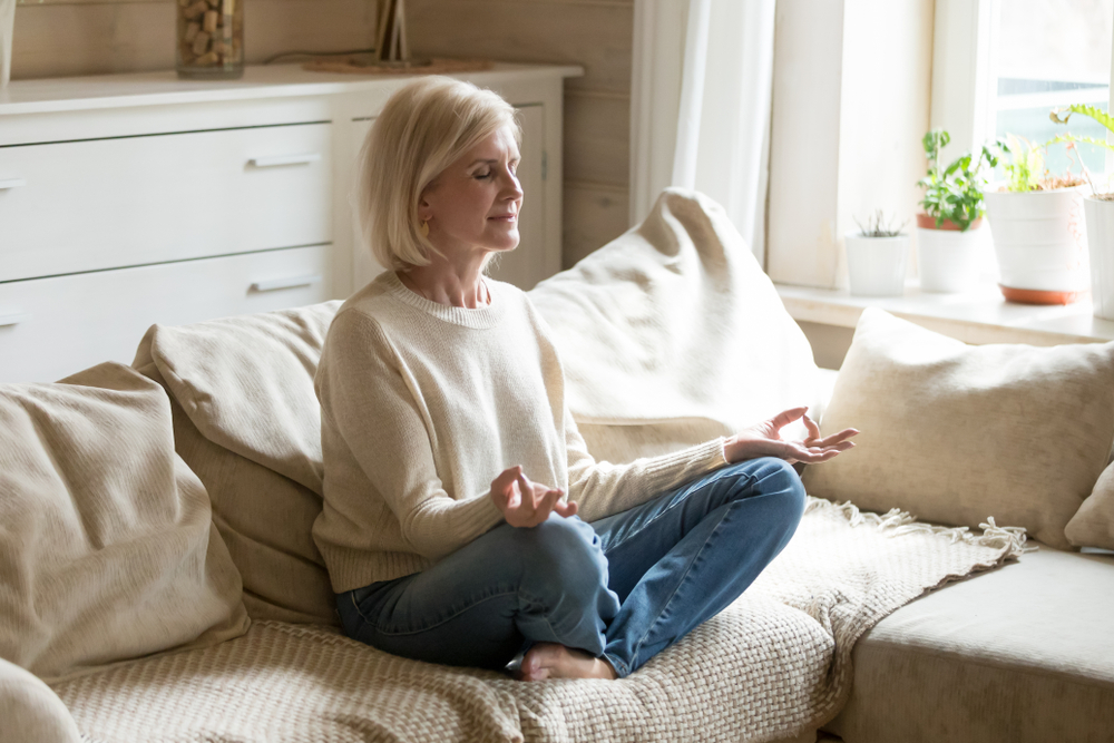 Женщина в возрасте медитирует сидя на диване дома.