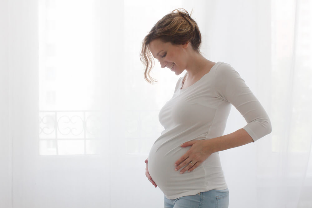 A pregnant woman is standing by the window, covered with a white curtain and caressing her stomach with great joy.