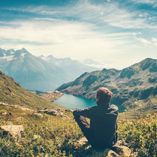 A traveler who is meditating by relaxing on a rock and admiring the magnificent view of the mountains and the lake.