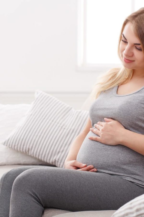 Eine schwangere Frau in grauer Kleidung sitzt zu Hause auf der Couch und streichelt mit beiden Händen ihren Bauch.