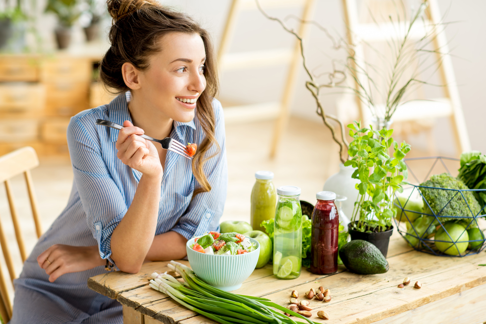 Una giovane felice sta mangiando un'insalata fatta con ingredienti sani mentre siede ad un tavolo in legno ricco di verdure, frutta e succo fresco appena spremuto.