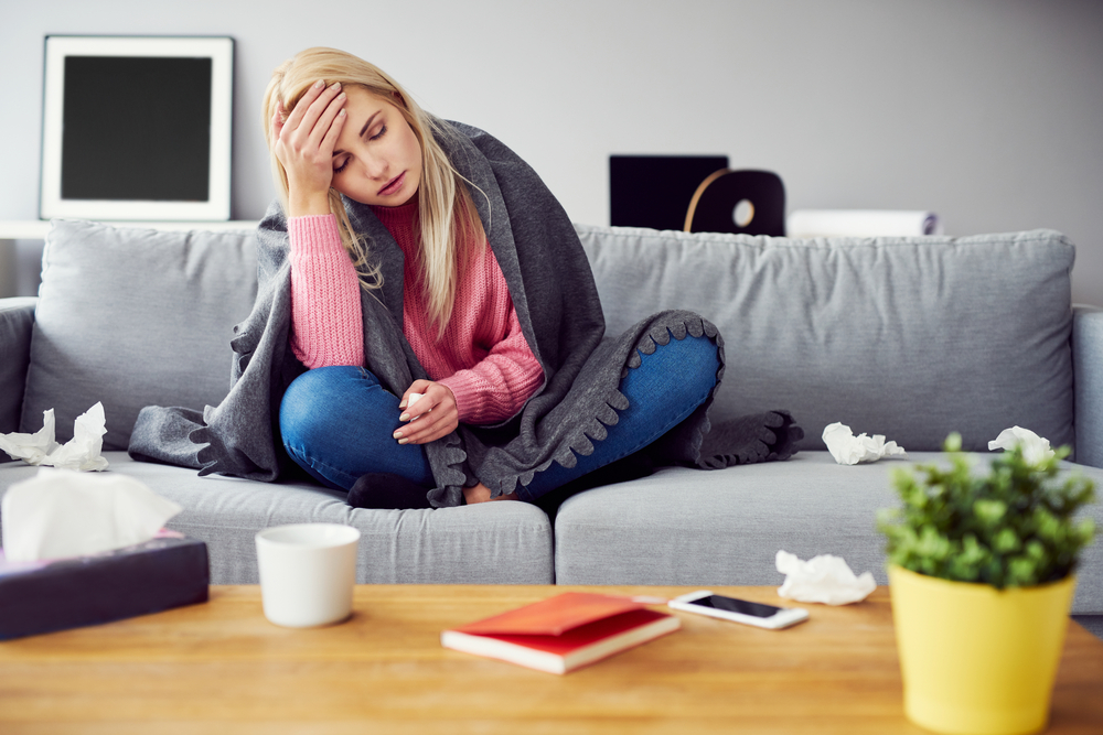 A woman with weak immune system is sitting on a couch and holding her head. There are many used tissues next to her.