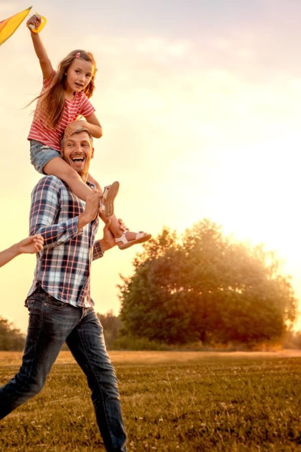 Eine glückliche Familie – Vater, Mutter und Tochter, die auf den Schultern ihres Vaters sitzt – lassen einen Drachen auf der Wiese in die Luft fliegen, während die Sonne am Himmel untergeht.