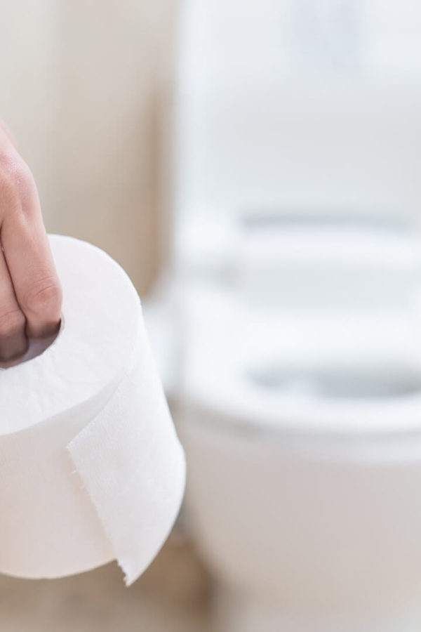 A woman holds a roll of toilet paper in her hand and stands next to a toilet bowl.