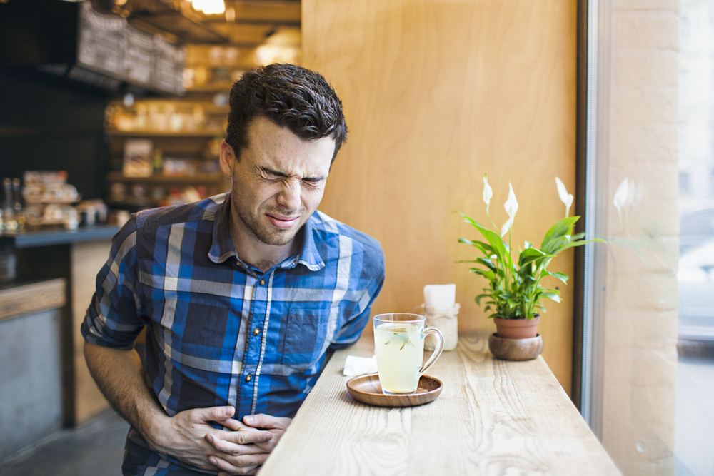 A man is sitting in a bar, drinking tea, and clutching his stomach because he has problems with his digestion.