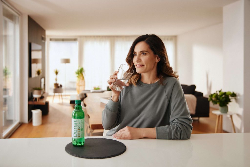 A woman is sitting at the kitchen counter and drinking a glass of Donat mineral water.