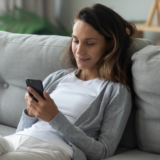 Una donna sorridente si riposa seduta confortevolmente sul divano in soggiorno e naviga in internet con uno smartphone.