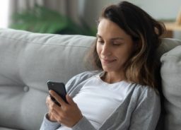 Eine lächelnde Frau ruht sich auf einer bequemen Couch im Wohnzimmer aus und surft auf einem modernen Smartphone im Internet.