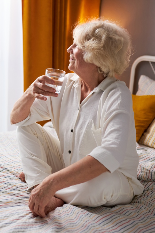 The older lady is sitting on the bed, drinking a glass of water and looking happily out the window.