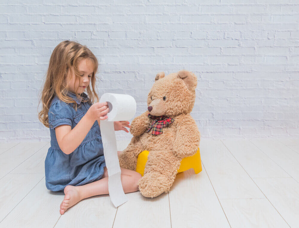A little girl is sitting on the floor and playing with a toilet paper roll, and there is a teddy-bear sitting on a potty next to her.